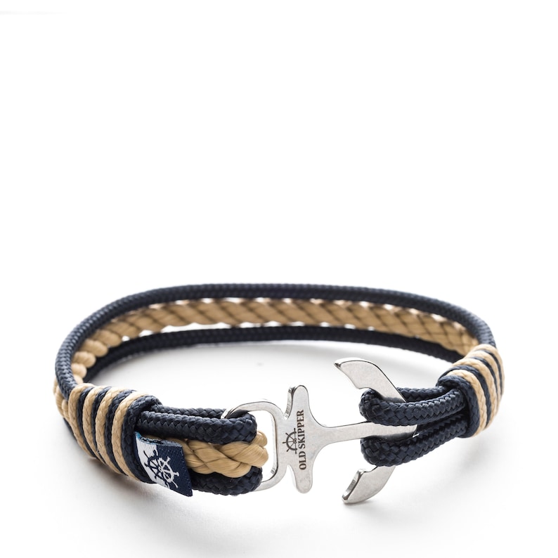 May include: A navy blue and beige rope bracelet with a silver anchor charm. The charm has the words "Old Skipper" engraved on it.