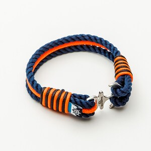 Könnte beinhalten: Ein blaues und orangefarbenes Seil-Armband mit einem silbernen Ankerverschluss. Das Armband hat ein weißes und blaues nautisches Rad-Design auf dem Seil.
