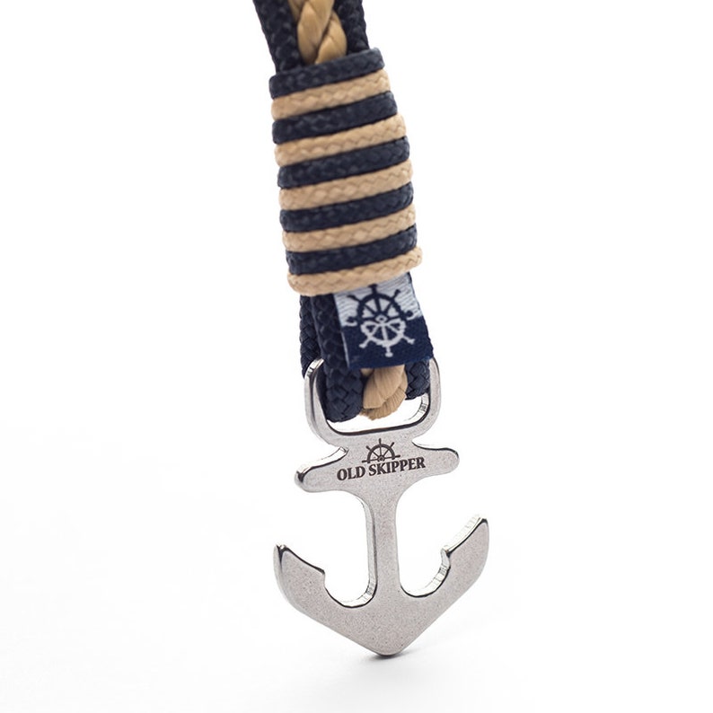 May include: A close-up of a navy blue and tan rope bracelet with a silver anchor clasp. The anchor has the words "Old Skipper" engraved on it.