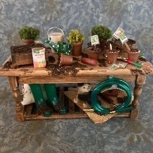 May include: A miniature wooden potting table with various gardening tools and accessories. The table holds small potted plants, a watering can, a bag of fertilizer, and a coiled green hose. Green rubber boots are stored below. The scene is set with soil.