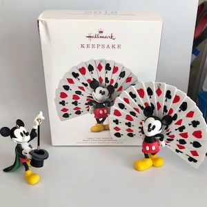 Op de afbeelding: Een Hallmark Keepsake 2018 Thru the Mirror Mickey's Movie Mouse-terpieces ornamentdoos met een Mickey Mouse-figuur die een waaier van speelkaarten vasthoudt, naast twee Mickey Mouse-beeldjes, één met een hoge hoed en één met een kaartenwaaier.