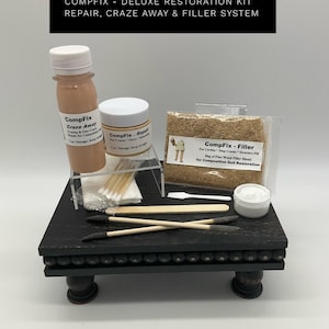 May include: A deluxe restoration kit for dolls, featuring bottles and jars of repair and filler compounds, applicators, and a bag of wood fiber blend. The kit is displayed on a black, decorative stand with small wooden feet. The text "COMPFIX - DELUXE RESTORATION KIT" is at the top.