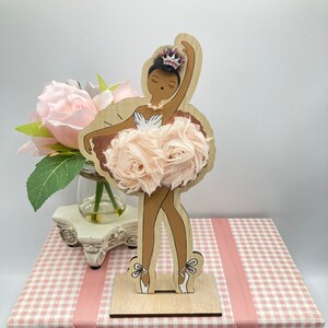 K&ouml;nnte beinhalten: Eine Holz-Ballerina-Figur mit hellbrauner Haut, die einen rosa T&uuml;llrock aus Stoffblumen und eine Schmuckkrone tr&auml;gt. Die Figur steht auf einem Holzsockel, und die Schachtel ist rosa und wei&szlig; kariert mit rosa Band.