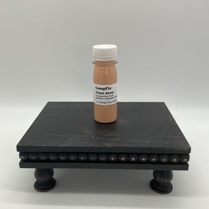 May include: A small bottle of CompFix Craze Away, a product for repairing crazing and fine cracks, sits on a dark wooden decorative stand. The stand has a square top, rounded edges, and decorative bead detailing.