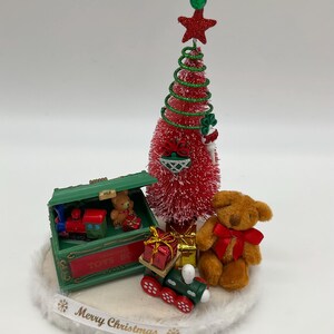 May include: A miniature Christmas scene with a red and green Christmas tree, a toy train, a teddy bear, and a toy chest with the words "Toys R Us" on the side. The scene is set on a white base with the words "Merry Christmas" written on it.