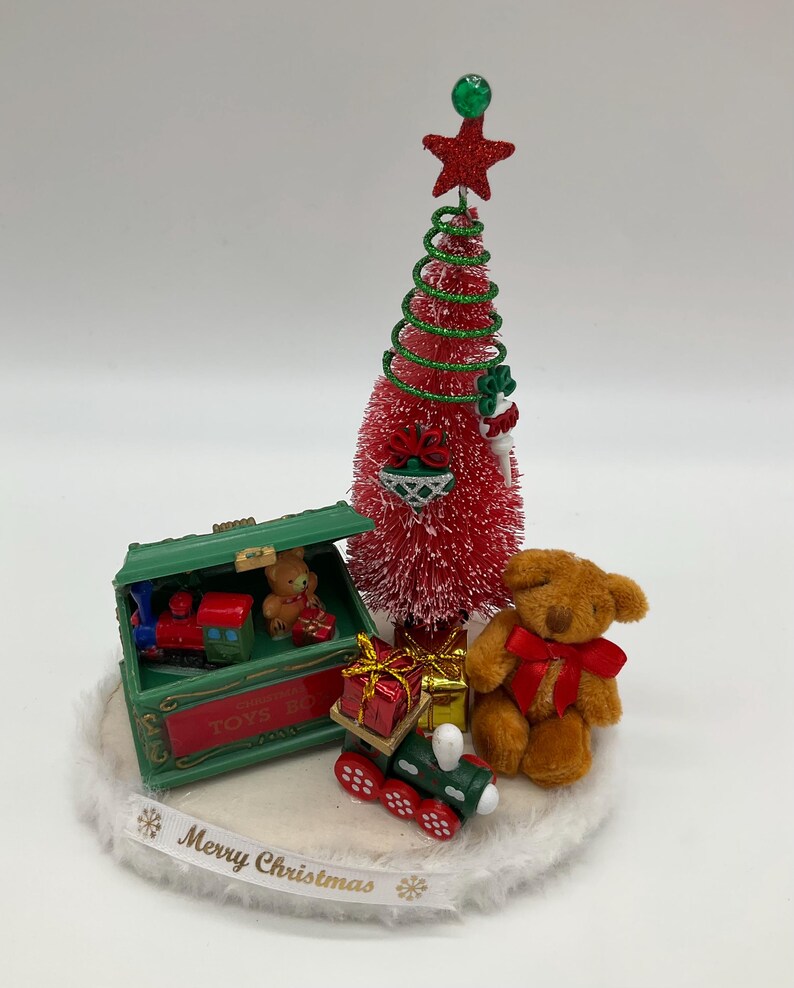 May include: A miniature Christmas scene with a red and green Christmas tree, a toy train, a teddy bear, and a toy chest with the words "Toys R Us" on the side. The scene is set on a white base with the words "Merry Christmas" written on it.