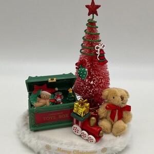 May include: A miniature Christmas scene with a red and green Christmas tree, a green toy box with the words "Christmas Toys Box", a teddy bear, a toy train, and a gift box. The scene is set on a white fluffy base with a gold ribbon that reads "Merry Christmas".