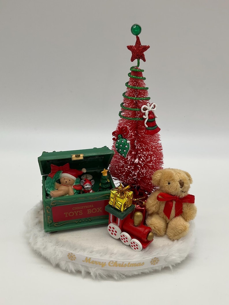 May include: A miniature Christmas scene with a red and green Christmas tree, a green toy box with the words "Christmas Toys Box", a teddy bear, a toy train, and a gift box. The scene is set on a white fluffy base with a gold ribbon that reads "Merry Christmas".