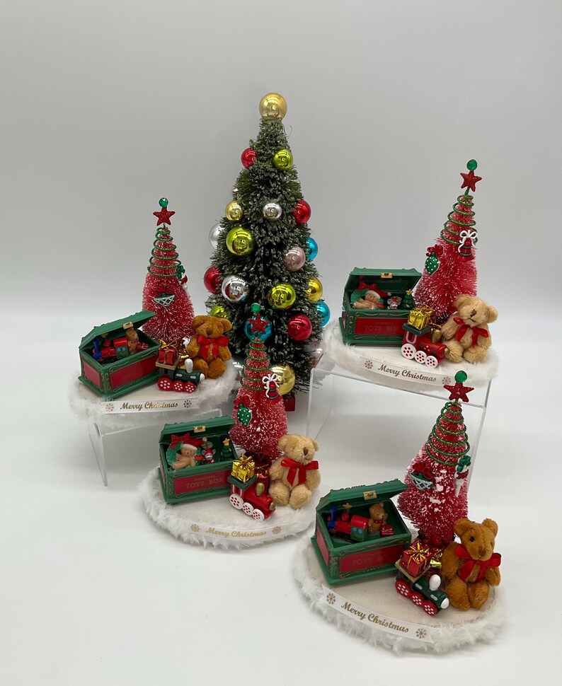 May include: Four miniature Christmas tree centerpieces with red and green toy chests, teddy bears, and the words "Merry Christmas".
