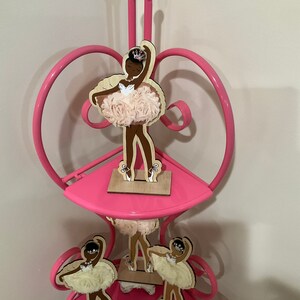 K&ouml;nnte beinhalten: Ein rosa Metallregal mit drei Ebenen, verziert mit Holz-Ballerina-Figuren. Jede Figur zeigt eine dunkelh&auml;utige T&auml;nzerin in einem Tutu mit einer Krone. Die Tutus sind aus einem hellrosa, strukturierten Stoff gefertigt.