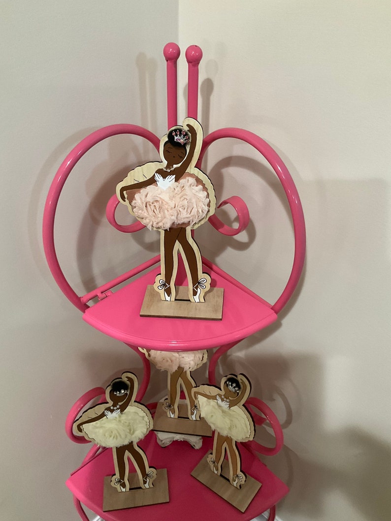 K&ouml;nnte beinhalten: Ein rosa Metallregal mit drei Ebenen, verziert mit Holz-Ballerina-Figuren. Jede Figur zeigt eine dunkelh&auml;utige T&auml;nzerin in einem Tutu mit einer Krone. Die Tutus sind aus einem hellrosa, strukturierten Stoff gefertigt.