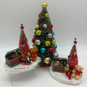 May include: Three miniature Christmas trees with red and green ornaments, a toy train, and teddy bears. The trees are on white bases with the words "Merry Christmas" printed on them.