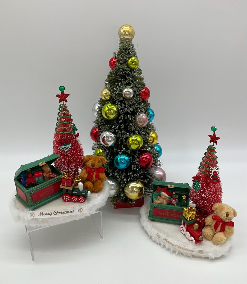 May include: Three miniature Christmas trees with red and green ornaments, a toy train, and teddy bears. The trees are on white bases with the words "Merry Christmas" printed on them.