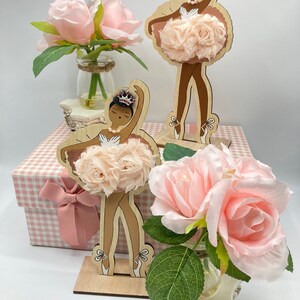 K&ouml;nnte beinhalten: Holzballerina-Figuren mit brauner Haut, die Kronen und Tutu-R&ouml;cke aus Stoff tragen. Die Figuren stehen auf Holzsockeln. Rosa Rosen in Gl&auml;sern und eine Geschenkbox mit einer rosa Schleife sind ebenfalls im Bild.