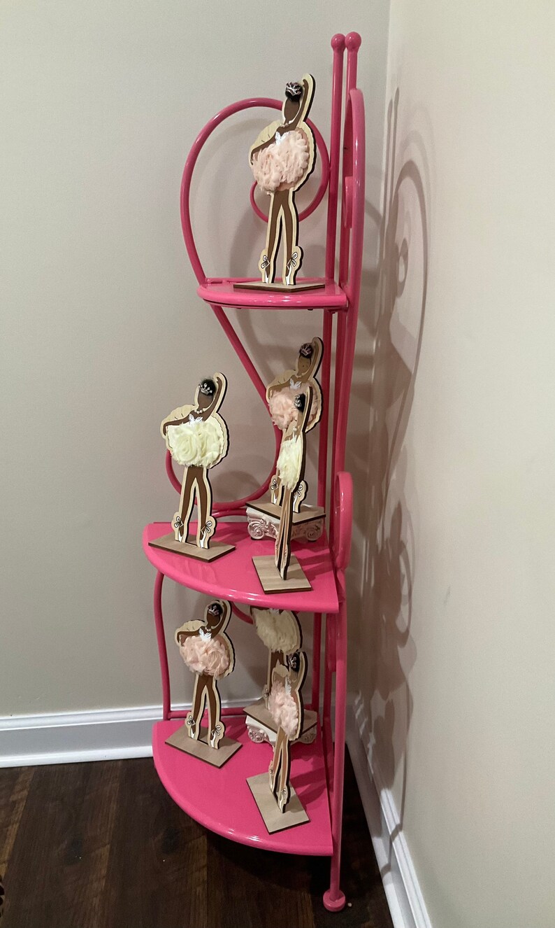 K&ouml;nnte beinhalten: Ein rosa Eckregal zeigt mehrere Holz-Ballerina-Figuren. Die Figuren haben braune K&ouml;rper, cremefarbene Tutus und sind auf kleinen Holzsockeln montiert. Das Regal hat ein herzf&ouml;rmiges Design.