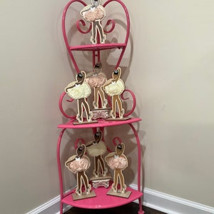 K&ouml;nnte beinhalten: Ein rosa Eckregal mit drei Ebenen und herzf&ouml;rmigen Akzenten. Das Regal zeigt mehrere Holz-Ballerina-Figuren mit cremefarbenen Tutus und schwarzen Haaren. Das Regal ist aus Metall und steht in einer Ecke.