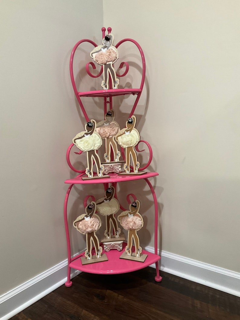 K&ouml;nnte beinhalten: Ein rosa Eckregal mit drei Ebenen und herzf&ouml;rmigen Akzenten. Das Regal zeigt mehrere Holz-Ballerina-Figuren mit cremefarbenen Tutus und schwarzen Haaren. Das Regal ist aus Metall und steht in einer Ecke.