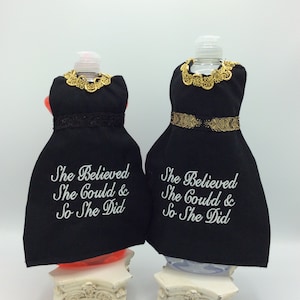 May include: Two black fabric bottle covers shaped like dresses, with gold trim around the neck and waist. The text "She Believed She Could & So She Did" is embroidered on the front. The bottles sit on decorative white pedestals.