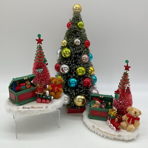 May include: A miniature Christmas scene with three small artificial Christmas trees decorated with ornaments. Each tree is on a white base with a green toy chest and a teddy bear. The bases have the text "Merry Christmas" on them.