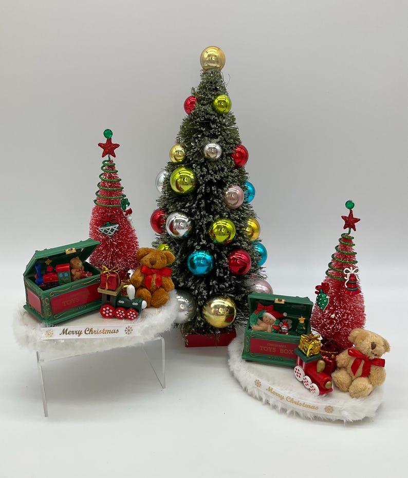 May include: A miniature Christmas scene with three small artificial Christmas trees decorated with ornaments. Each tree is on a white base with a green toy chest and a teddy bear. The bases have the text "Merry Christmas" on them.