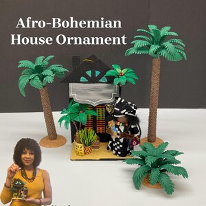 May include: A black and white doll with a colorful patterned robe stands in front of a miniature house with a black roof and a green palm tree. The house has a sign that says "Afro-Bohemian House Ornament". The doll is holding a small, colorful object in its hand. The house is surrounded by green plants and palm trees.