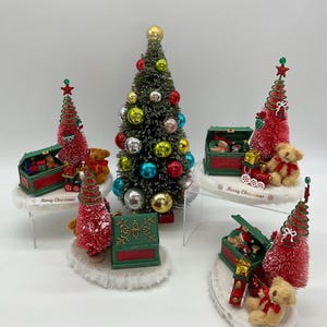 May include: Five miniature Christmas trees with red and green ornaments, teddy bears, and toy chests. The trees are decorated with red and white snow. The chests are open and filled with toys. The bases of the trees are decorated with the words "Merry Christmas".
