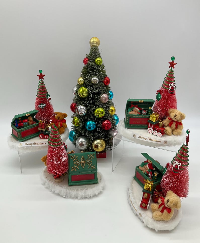 May include: Five miniature Christmas trees with red and green ornaments, teddy bears, and toy chests. The trees are decorated with red and white snow. The chests are open and filled with toys. The bases of the trees are decorated with the words "Merry Christmas".