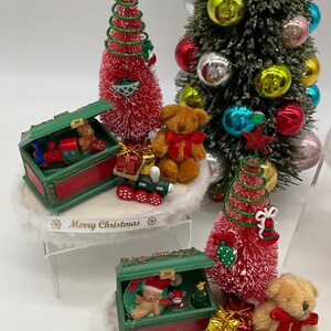 May include: Three miniature Christmas trees with red and green brush, each with a green toy chest filled with miniature toys, a teddy bear, and a train. The base of each tree is decorated with white faux fur and a ribbon that reads "Merry Christmas".