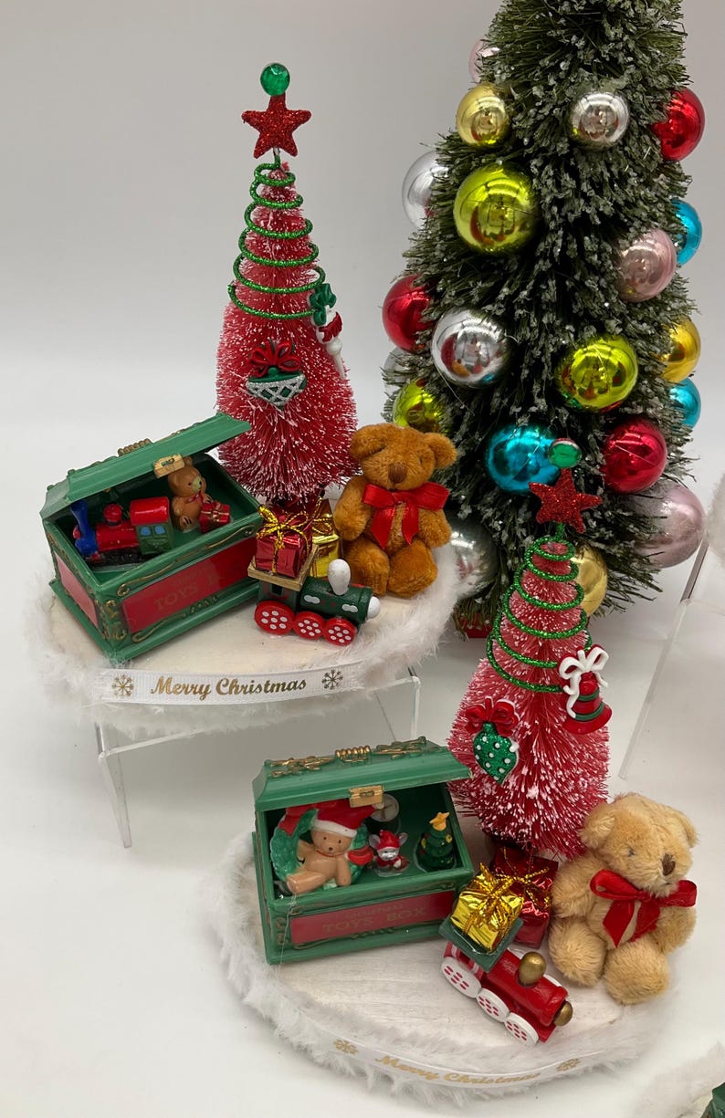 May include: Three miniature Christmas trees with red and green brush, each with a green toy chest filled with miniature toys, a teddy bear, and a train. The base of each tree is decorated with white faux fur and a ribbon that reads "Merry Christmas".