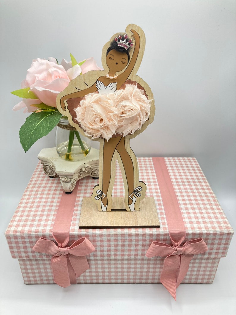 K&ouml;nnte beinhalten: Eine Holz-Ballerina-Figur mit hellbrauner Haut, die einen rosa T&uuml;llrock aus Stoffblumen und eine Schmuckkrone tr&auml;gt. Die Figur steht auf einem Holzsockel, und die Schachtel ist rosa und wei&szlig; kariert mit rosa Band.