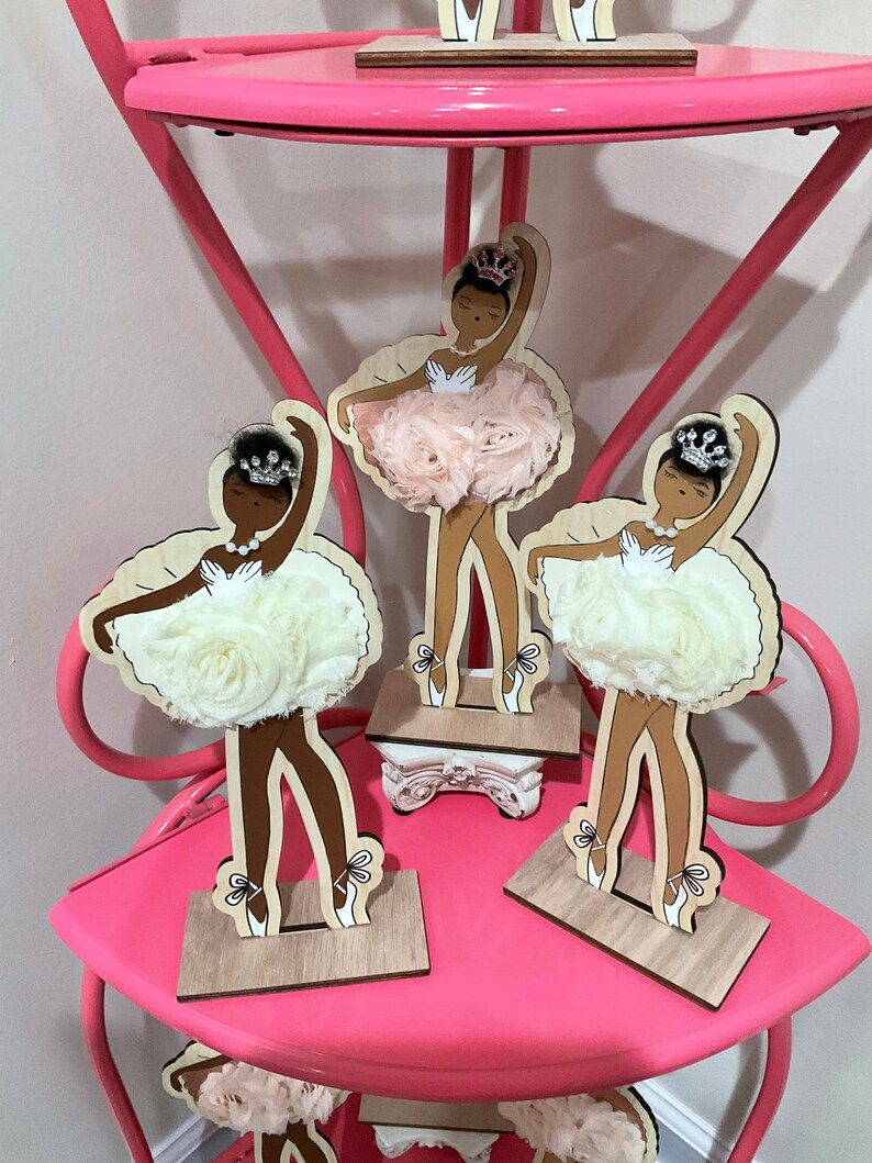 K&ouml;nnte beinhalten: Holzballerina-Figuren mit brauner Haut, die Kronen und Tutus aus Stoffblumen tragen. Die Figuren stehen auf einem rosa Regal. Die Tutus sind wei&szlig; und hellrosa. Die Ballerinas posieren in verschiedenen Balletthaltungen.