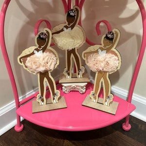 K&ouml;nnte beinhalten: Drei Holzfiguren von Ballerinas mit brauner Haut, die Kronen und Tutus aus Stoff tragen. Die Figuren stehen auf einem rosa Regal mit einem rosa Metallrahmen.