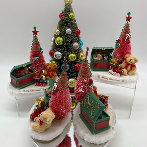 May include: A set of five miniature Christmas trees with red and green ornaments, teddy bears, and toy trains. Each tree is in a green and red box with a white base. The boxes have the words "Merry Christmas" on them.