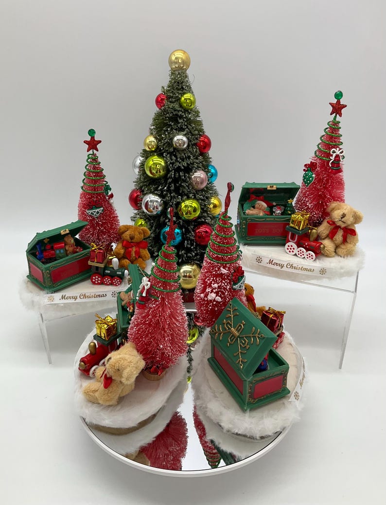 May include: A set of five miniature Christmas trees with red and green ornaments, teddy bears, and toy trains. Each tree is in a green and red box with a white base. The boxes have the words "Merry Christmas" on them.