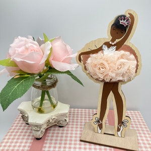 K&ouml;nnte beinhalten: Holz-Ballerina-Figur mit hellbrauner Haut, die einen rosa T&uuml;t&uuml; und eine Schmuckkrone tr&auml;gt. Die Figur steht auf einem Holzsockel. Ein kleines Glas mit rosa Rosen steht auf einem dekorativen wei&szlig;en St&auml;nder.