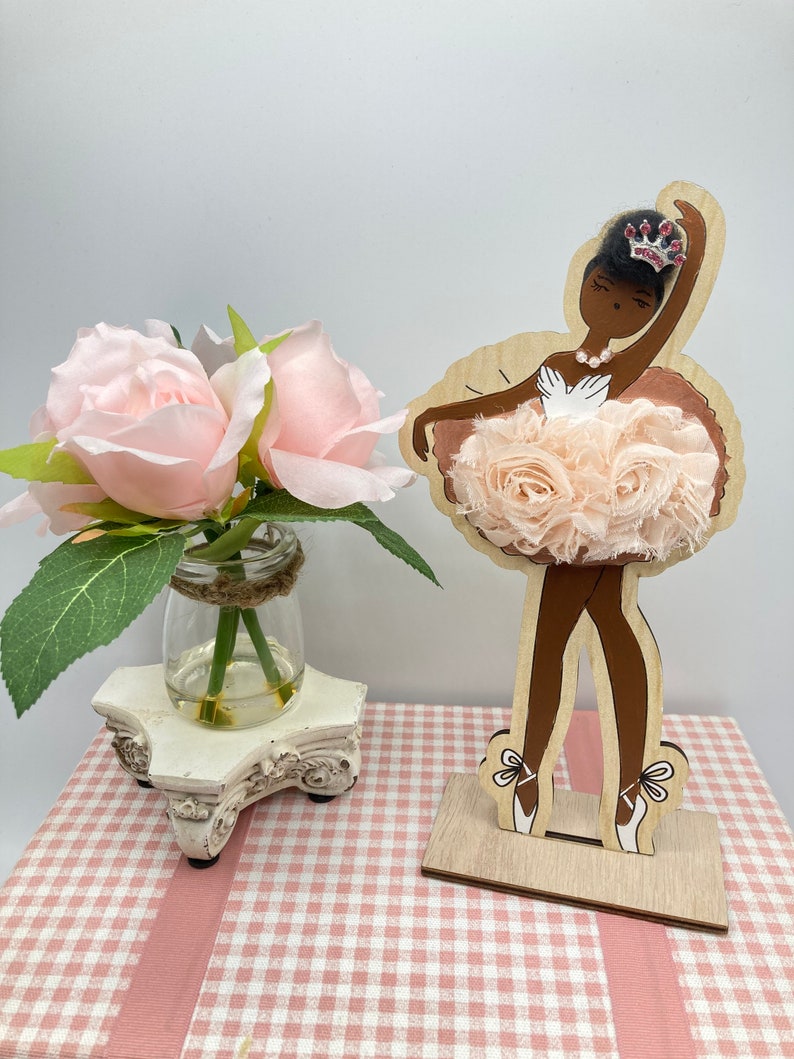 K&ouml;nnte beinhalten: Holz-Ballerina-Figur mit hellbrauner Haut, die einen rosa T&uuml;t&uuml; und eine Schmuckkrone tr&auml;gt. Die Figur steht auf einem Holzsockel. Ein kleines Glas mit rosa Rosen steht auf einem dekorativen wei&szlig;en St&auml;nder.