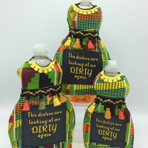 May include: Dish soap bottles dressed in colorful fabric covers with yellow, orange, and green accents. Each cover has a black tag with the embroidered phrase "The dishes are looking at me DIRTY again."