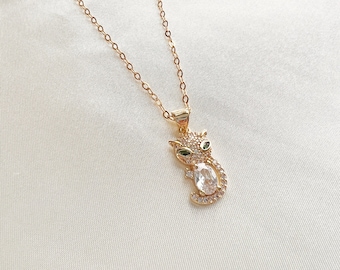 May include: A gold chain necklace with a small, clear crystal cat pendant. The cat has green eyes and is set with smaller clear crystals.