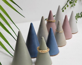 May include: A set of nine concrete ring holders in the shape of cones. The cones are in shades of gray, blue, and pink. Some of the cones have gold rings on them.