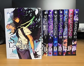 Puede incluir: Una colección de volúmenes de manga Solo Leveling. El primer volumen está abierto, mostrando una ilustración de personaje y el título. Las lomos de los volúmenes 7 al 13 son visibles, cada uno con el título de la serie y el número de volumen.