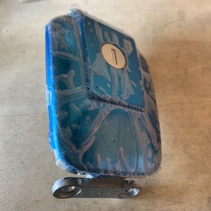 May include: A blue plastic gas cap with a silver keyhole and the number "1" on it. The cap is attached to a silver metal hinge.