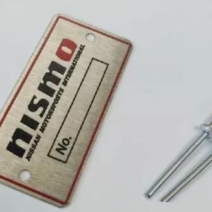May include: A silver metal plaque with the text "nismo NISSAN MOTORSPORTS INTERNATIONAL" and "No." printed in black. The plaque has a red border and is designed for a number to be added.
