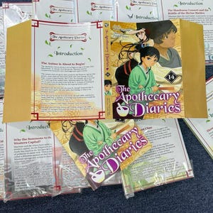 May include: A collection of The Apothecary Diaries manga volume 14, featuring a colorful illustrated cover with two characters, the title in purple and white text, and several informational inserts with printed text about the series.