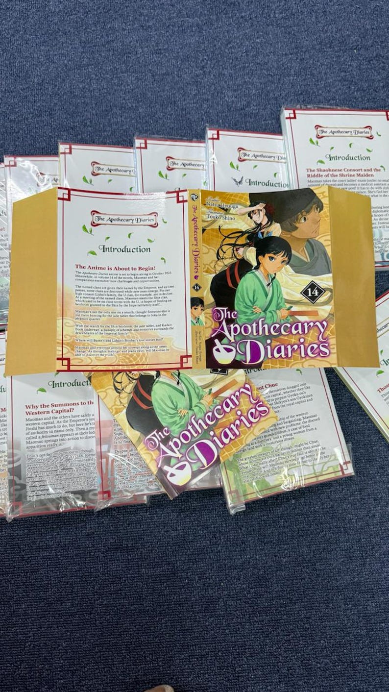 May include: A collection of The Apothecary Diaries manga volume 14, featuring a colorful illustrated cover with two characters, the title in purple and white text, and several informational inserts with printed text about the series.