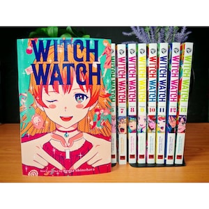 May include: A set of manga volumes titled "Witch Watch" with vibrant covers showcasing anime-style artwork. The initial volume displays a girl winking and making a heart shape. Volumes 6 to 13 are visible, presented in a black book stand.