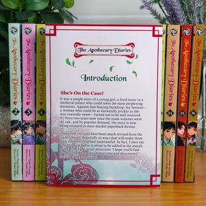 May include: A collection of The Apothecary Diaries paperback books arranged in a row. The central book is open to an introduction page titled She's On the Case! with red border accents. The spines show various volume numbers and character art.
