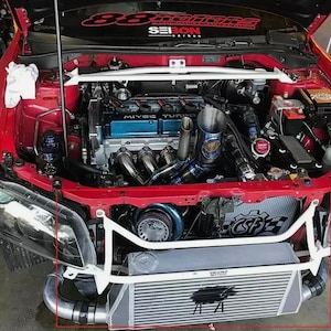 May include: A red car engine bay with the hood open, showcasing a modified engine. The engine features blue and silver components, including a turbocharger and intercooler. The car has a white brace bar and a banner that reads "88 Motors".