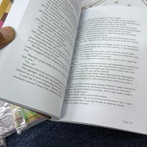 May include: An open book titled The Apothecary Diaries with white pages and black printed text. The left page shows dialogue between characters, while the right page features a header with the title and continues the narrative text.