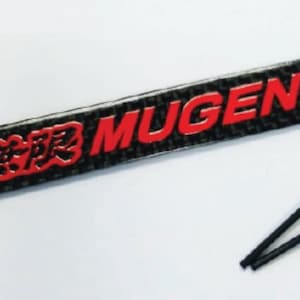 Könnte beinhalten: Ein schwarzes und rotes Auto-Emblem mit dem Wort "MUGEN" in roten Buchstaben und japanischen Schriftzeichen. Das Emblem ist aus Kohlefaser gefertigt und wird mit zwei schwarzen Druckknöpfen geliefert.