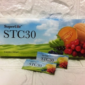 May include: A box of SuperLife STC30 dietary supplement sachets with a blue, green, and white background featuring a variety of fruits and vegetables. The box contains 15 sachets, each containing 1500mg of the supplement. Two individual sachets are shown in the foreground.